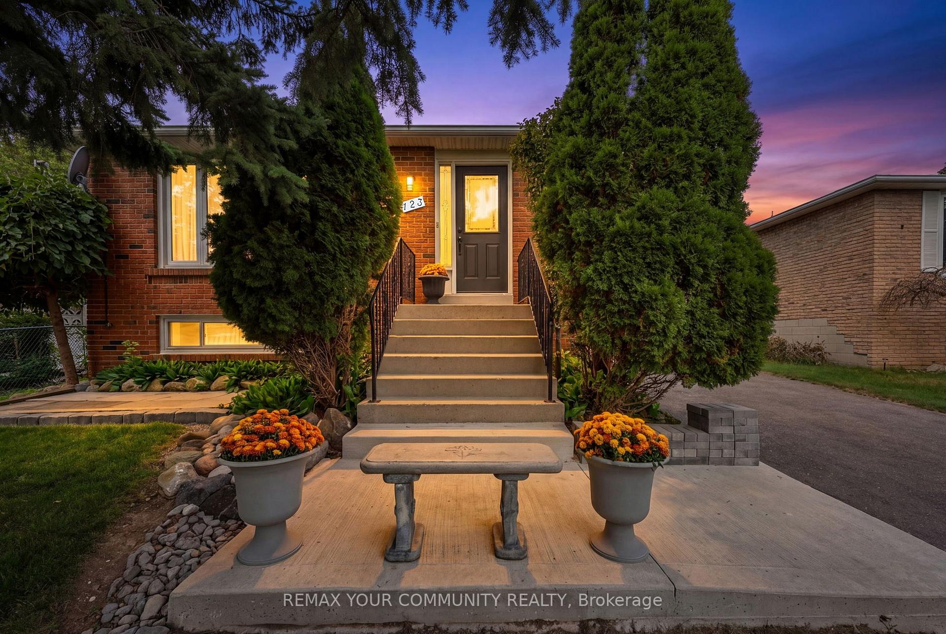 Red-brick home entrance at dusk with broad steps, tall evergreens, and flower planters.