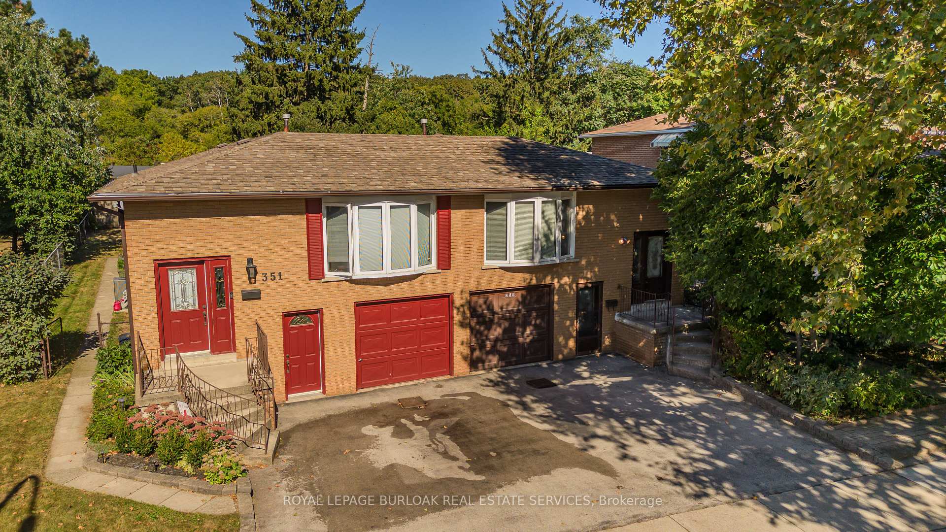 Brick two-level house with red doors and garage doors, viewed from above among mature trees.