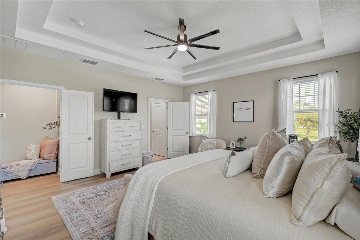 Furnished bedroom with a large white bed, tray ceiling, ceiling fan, and tall curtained windows.