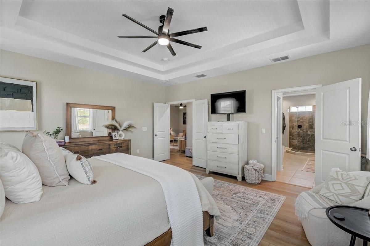 Bedroom with a white bed, tray ceiling, dark ceiling fan, and doorway to a tiled shower.
