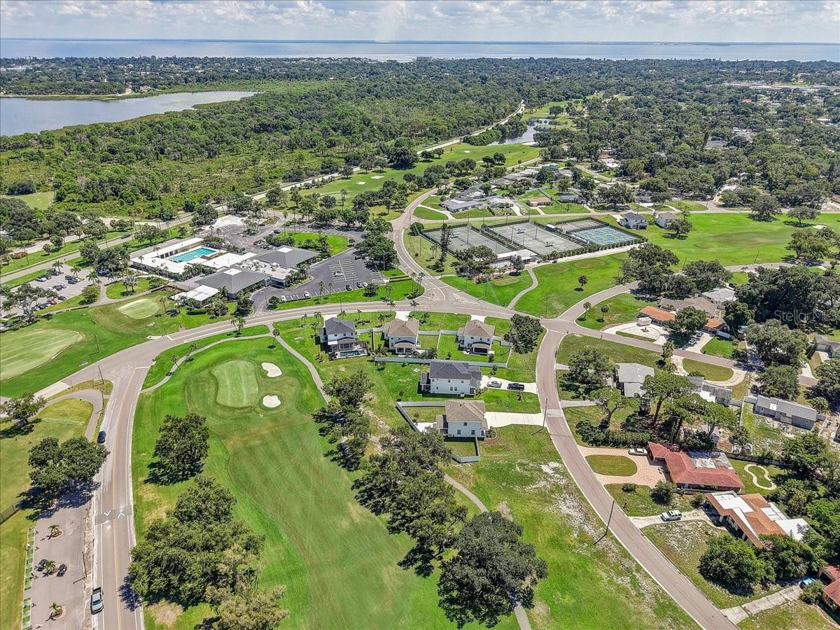Aerial neighborhood view with golf course, tennis courts, pool, and water in the distance.
