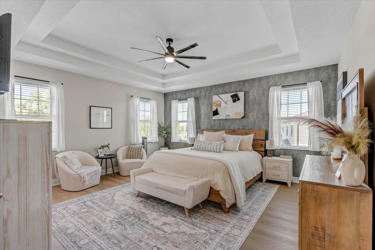 Staged bedroom with a king-size bed, textured gray accent wall, tray ceiling, and upholstered seating.