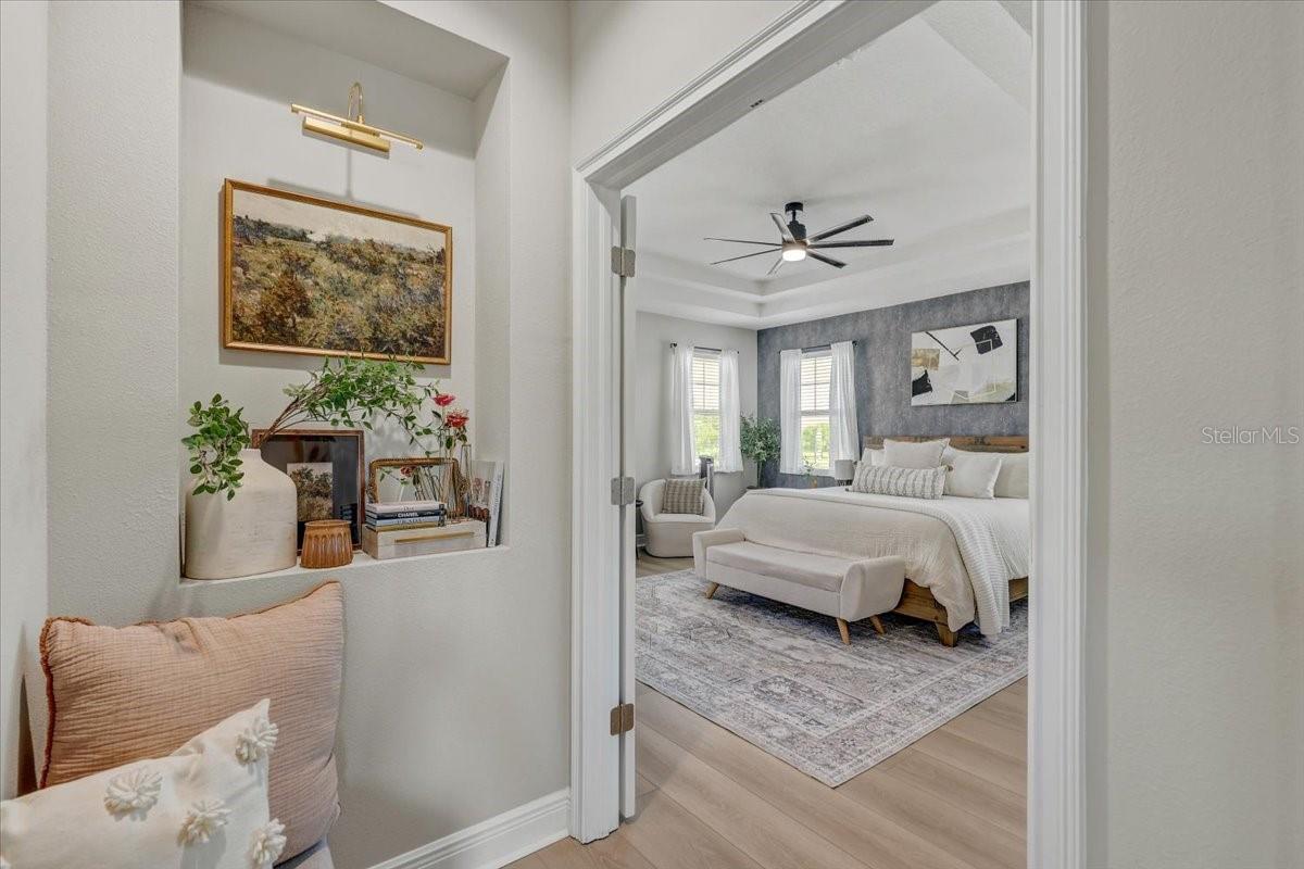 Furnished bedroom with a white bed, gray accent wall, and ceiling fan, viewed through an open doorway.