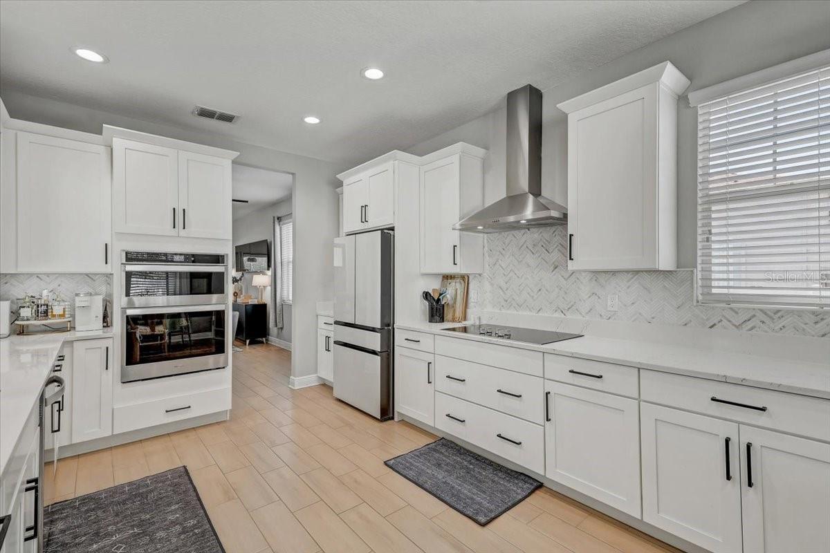 Bright white kitchen with stainless steel appliances, herringbone backsplash, and light wood-look floors.