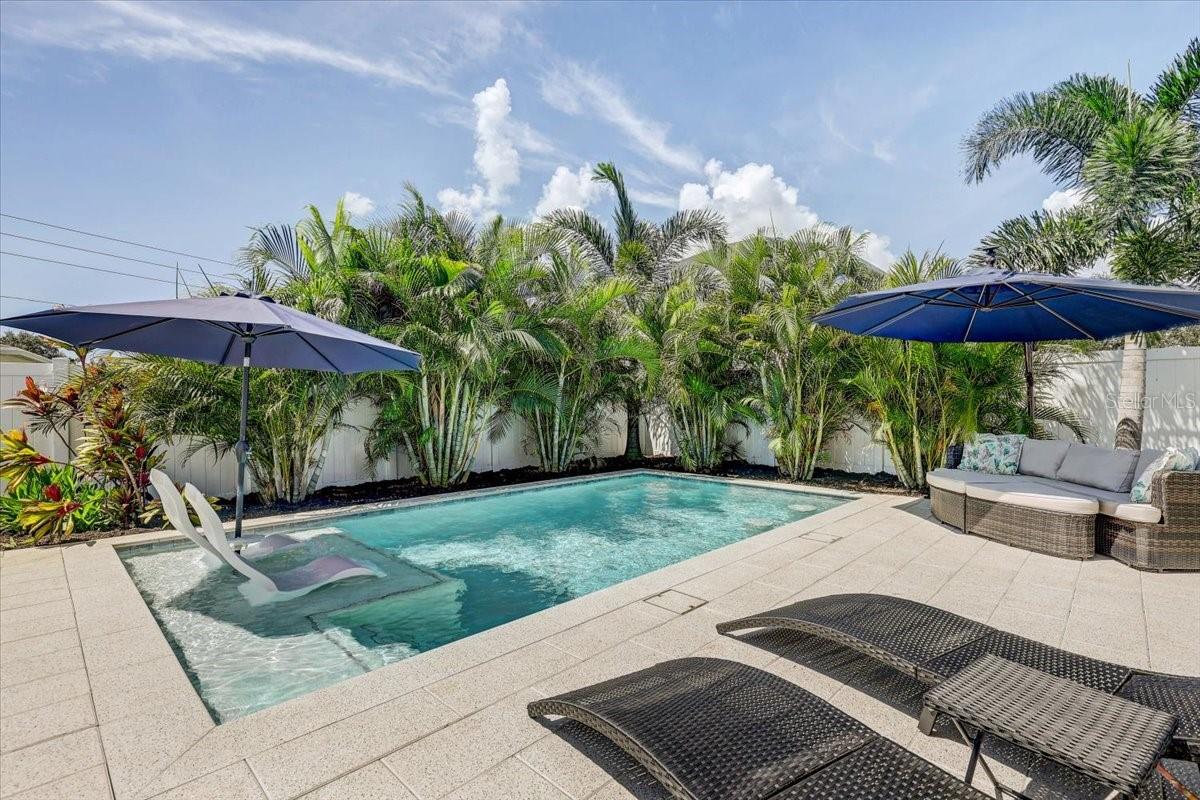 Backyard pool with turquoise water, umbrella-shaded seating, loungers, and tropical palms.