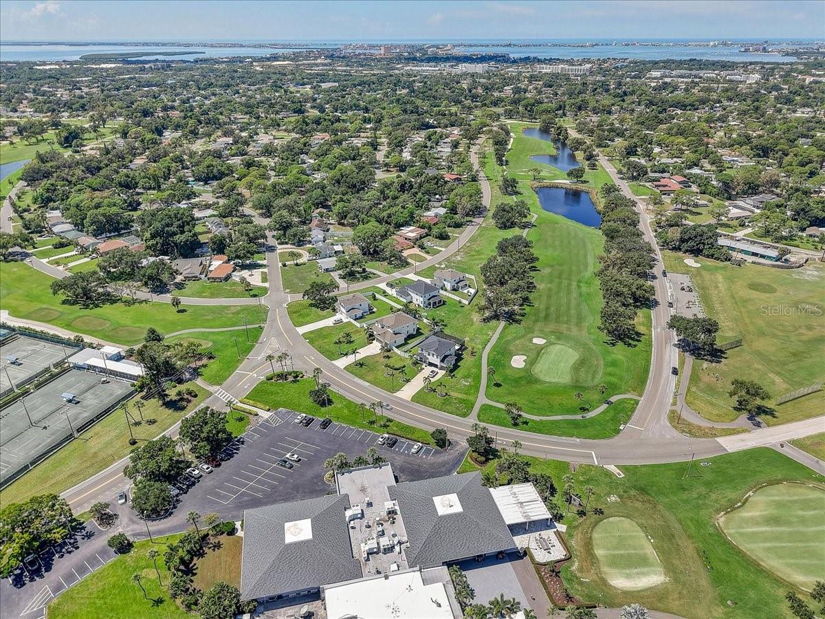 Aerial view of homes beside a golf course with ponds, tennis courts, and bay water in the distance.