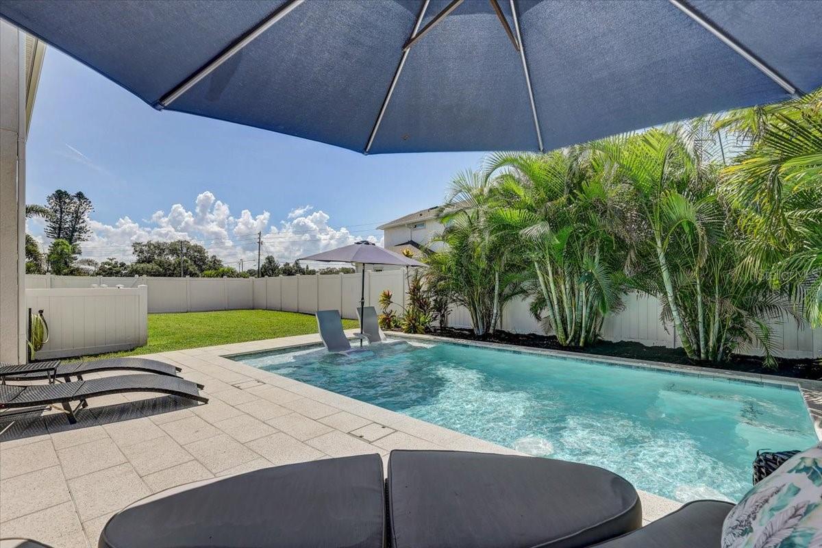 Backyard pool and stone patio with a blue umbrella, lounge chairs, and tropical palms.