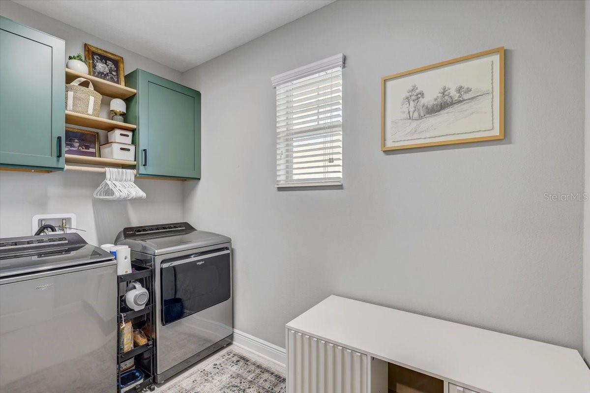 Laundry room with gray washer and dryer, teal cabinets, a window, and a white storage bench.