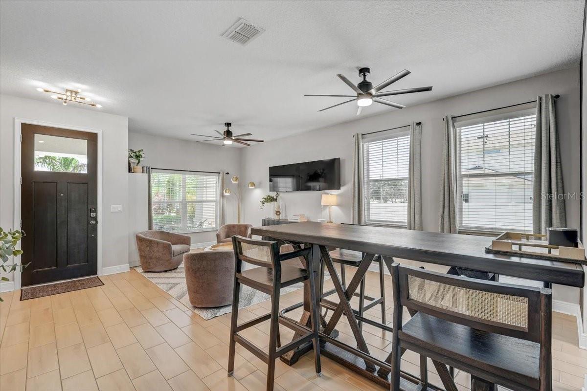 Furnished dining and living area with a long dark table, large windows, and a dark front door.