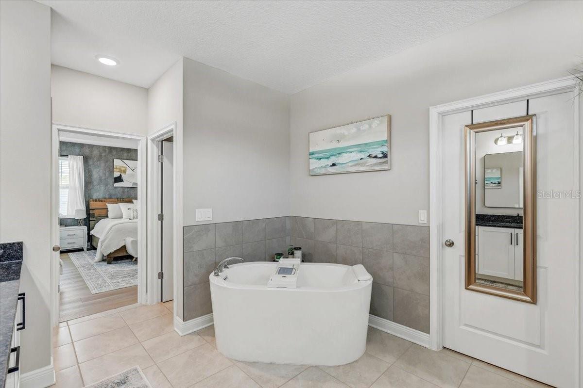 Bathroom with a freestanding soaking tub and gray tile wainscoting beside an open bedroom doorway.