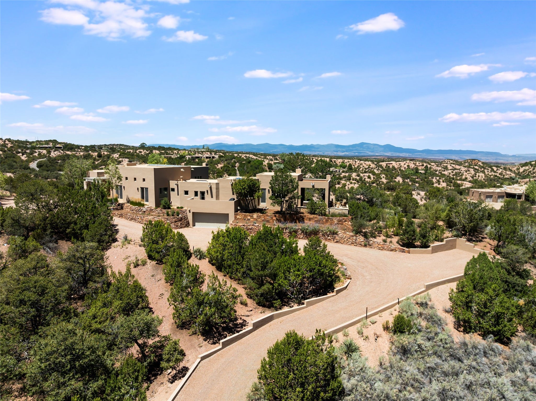 Modern adobe-style home with a curving gravel drive, desert vegetation, and distant mountains.