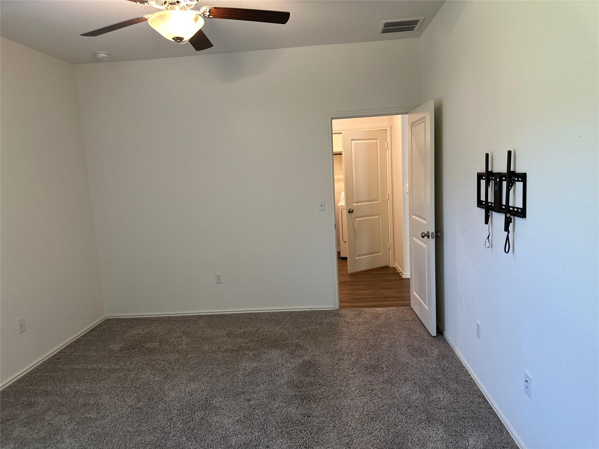 Empty carpeted bedroom with a ceiling fan, TV wall mount, and open doorway to the hall.