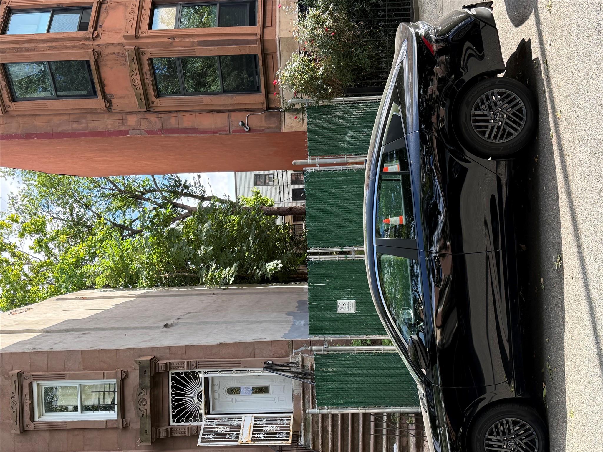 Brooklyn brownstone facades and a fenced lot behind a parked black car.