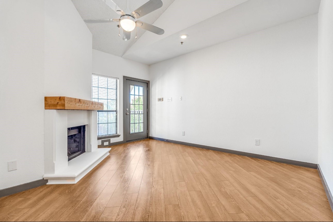 Empty living room with a wood-mantel fireplace, plank flooring, and glass-paneled door.
