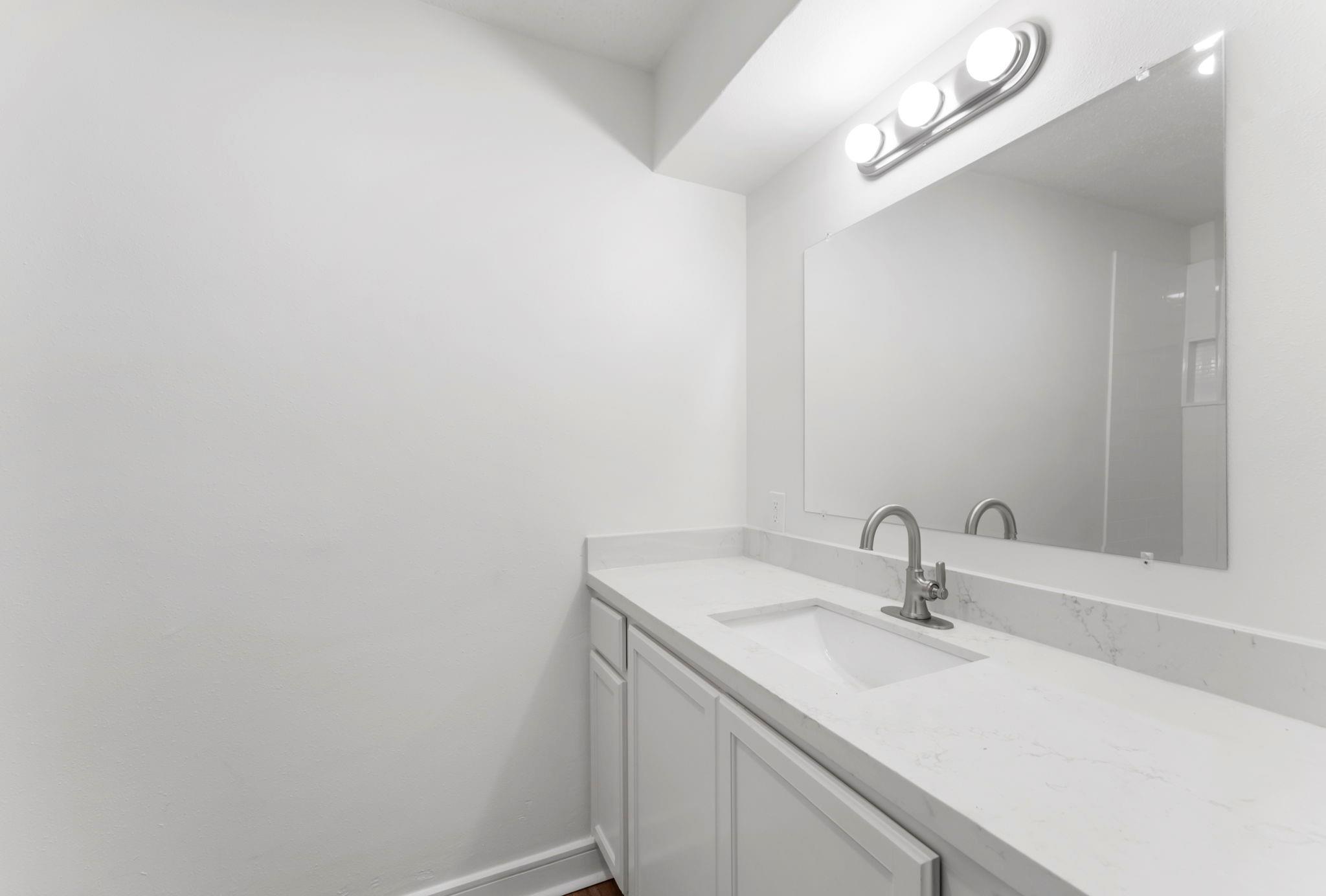 Bathroom vanity with a white sink, brushed metal faucet, large mirror, and three-bulb light.