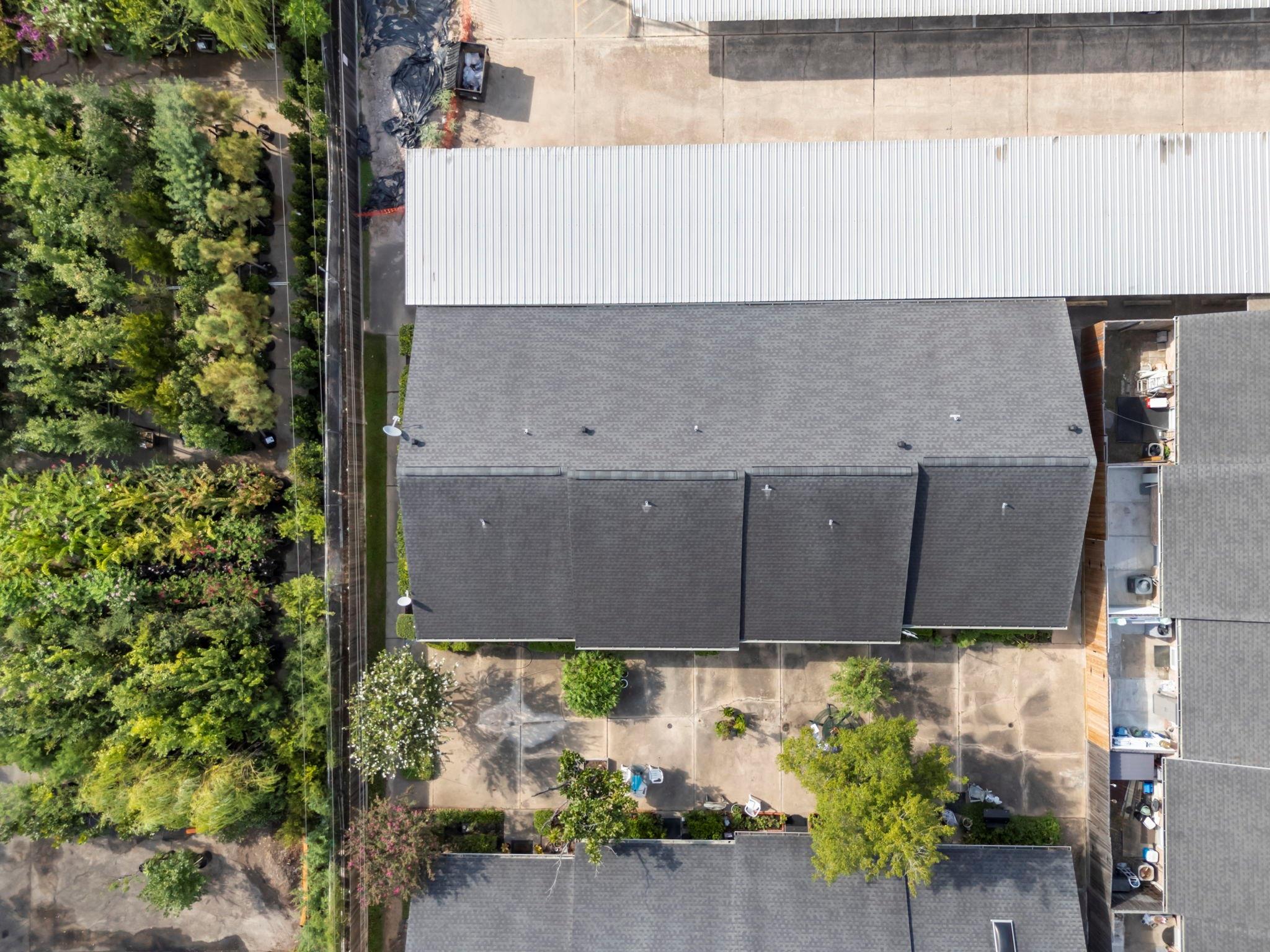 Aerial view of attached homes with dark roofs, a paved courtyard, and mature trees.