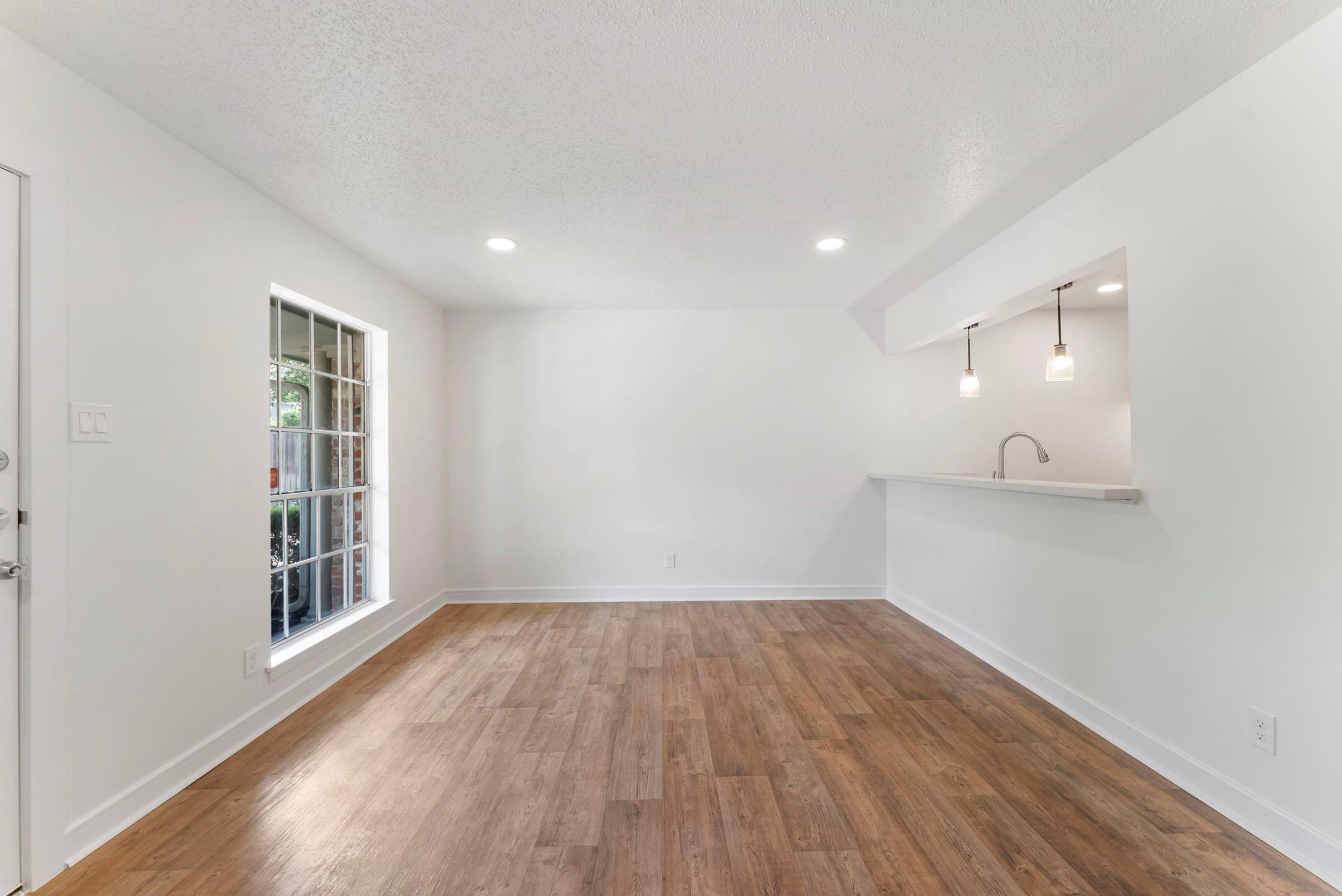 Vacant living area with wood-look floors, a tall grid-pane window, and a kitchen pass-through.