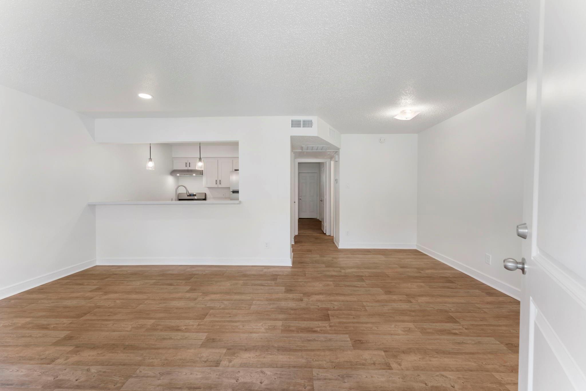 Empty living area with wood-look flooring, kitchen pass-through, and hallway.