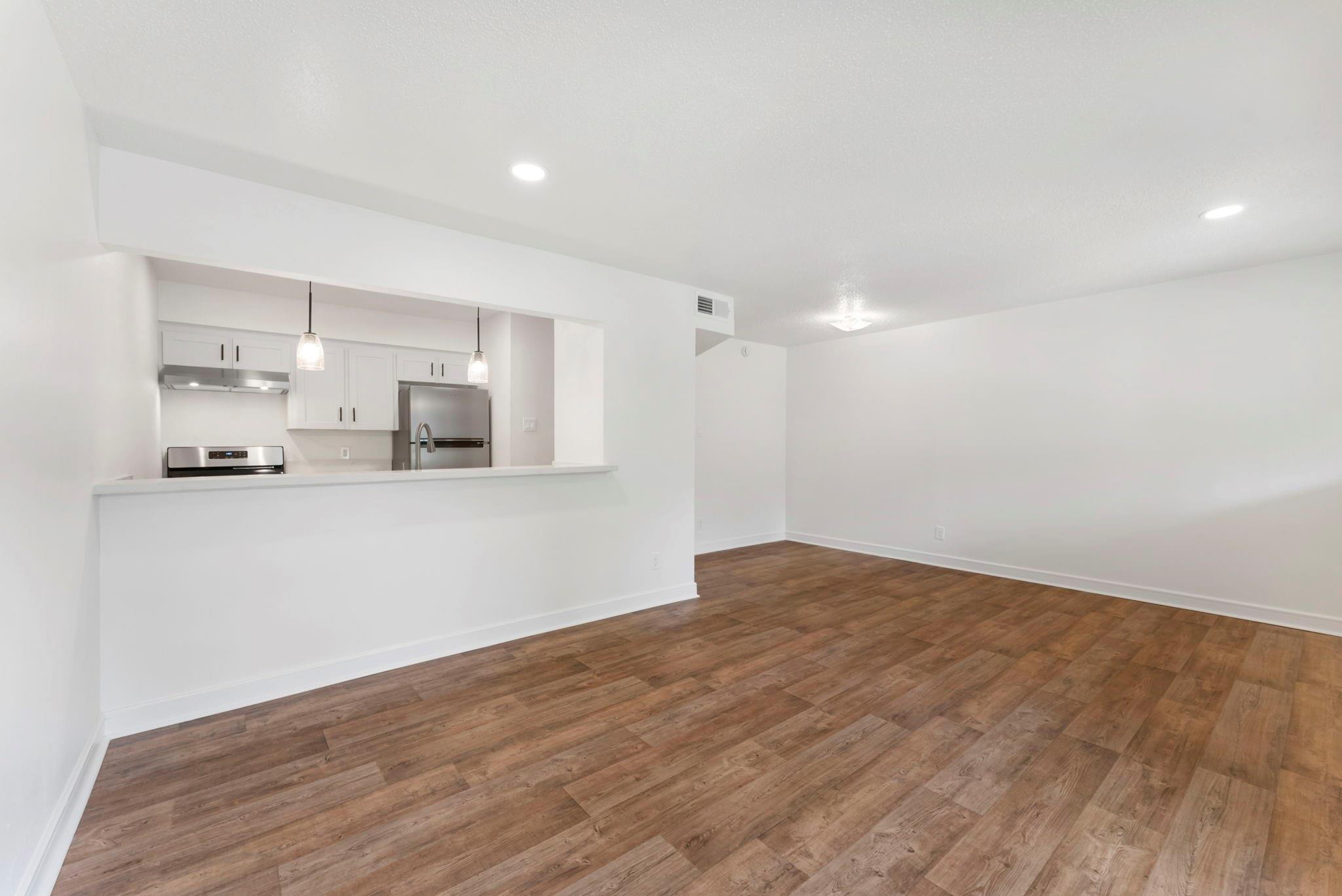 Empty living area with wood-look floors and an open kitchen with white cabinets and stainless steel appliances.