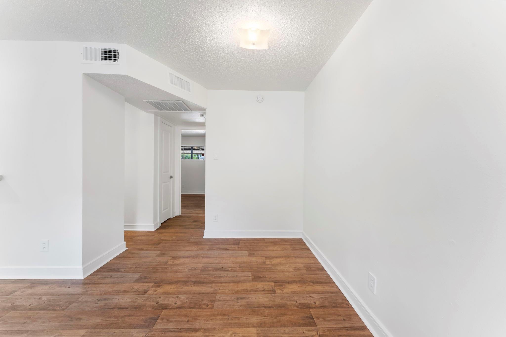 Vacant room with white walls, brown wood-look flooring, and an open doorway to a hall.