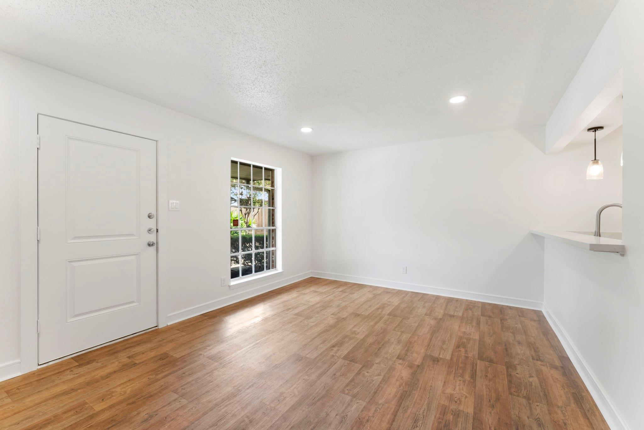 Vacant living area with wood-look flooring, entry door, large window, and kitchen breakfast bar.