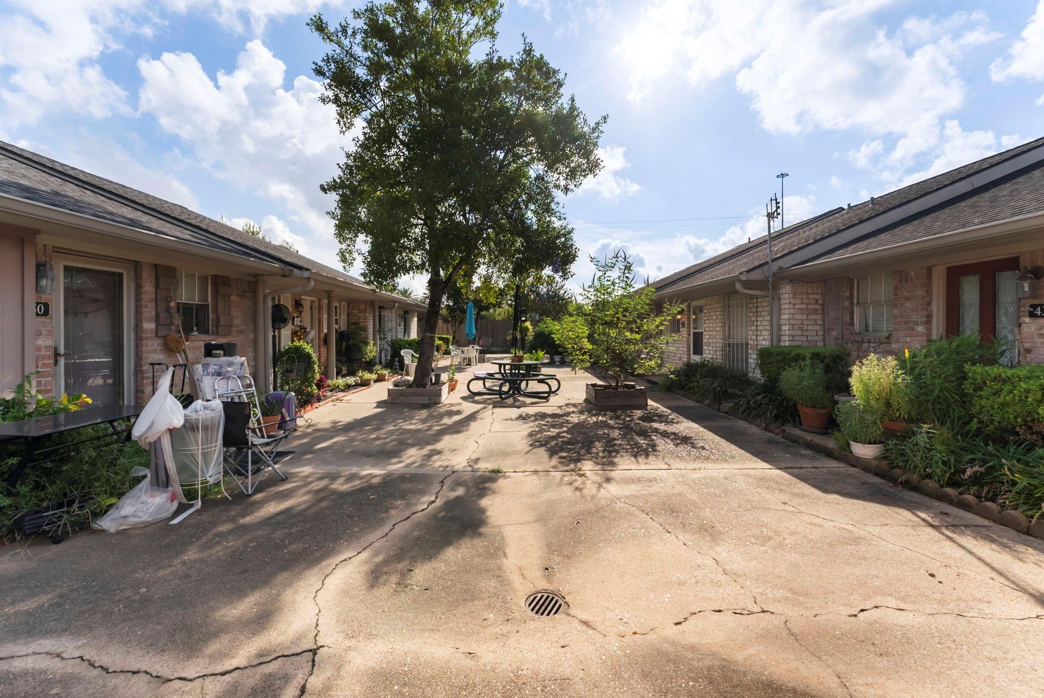 Shared courtyard between brick residences, with a large tree, picnic table, and potted plants.