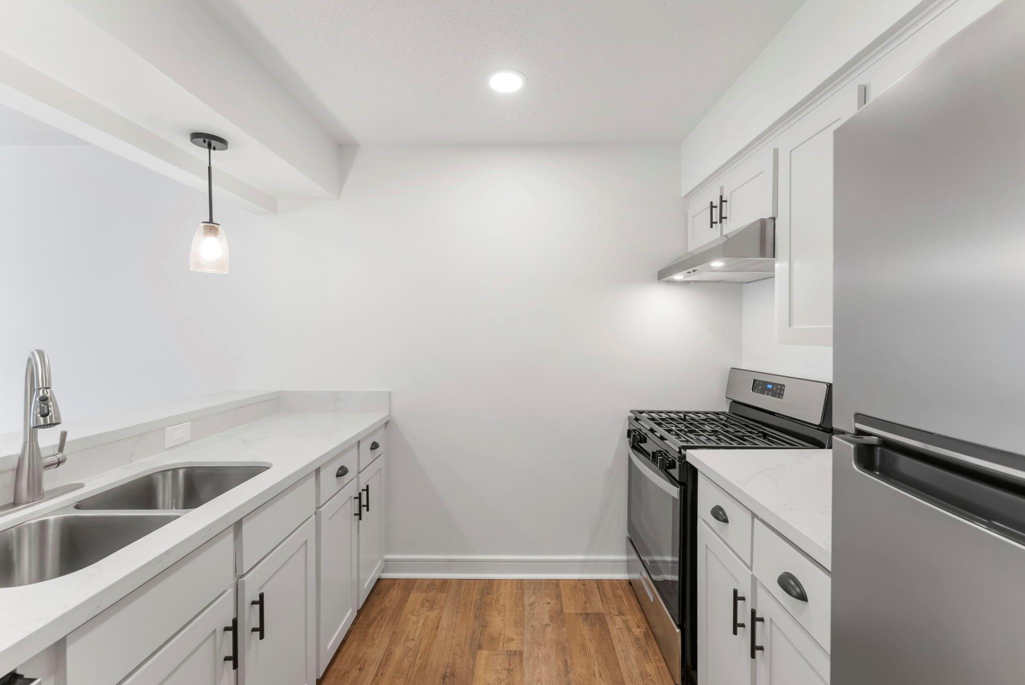 Galley kitchen with white cabinets, veined countertops, stainless appliances, and wood-look flooring.