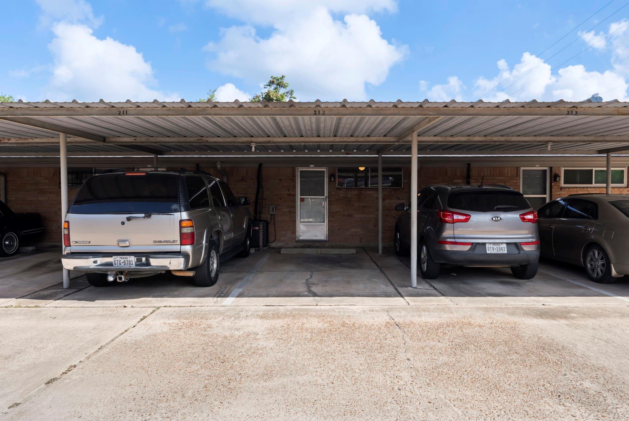 Numbered parking spaces beneath a metal carport in front of a brick residential building.