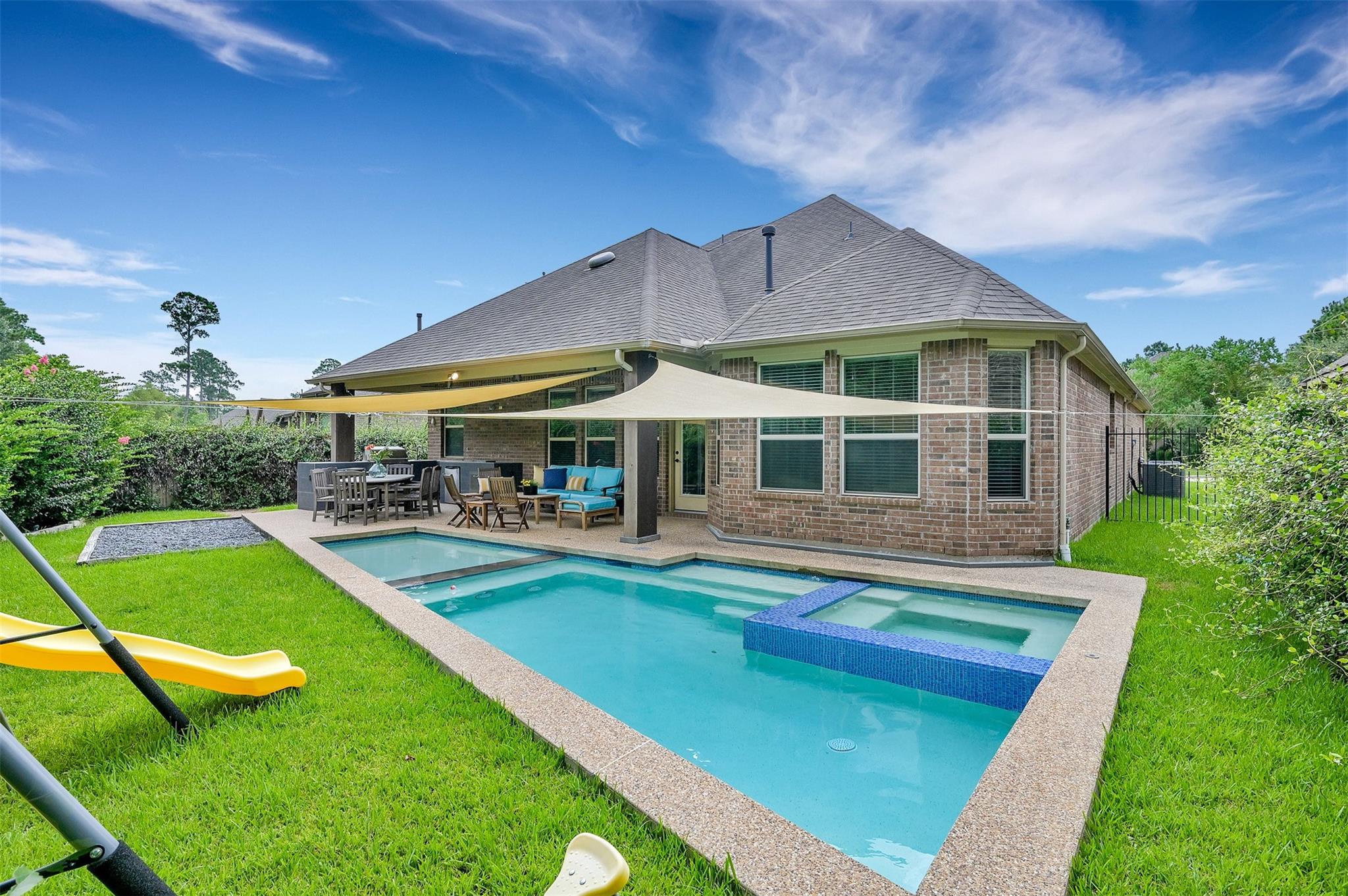Backyard pool and spa beside a brick house, with a shade sail over the furnished patio.
