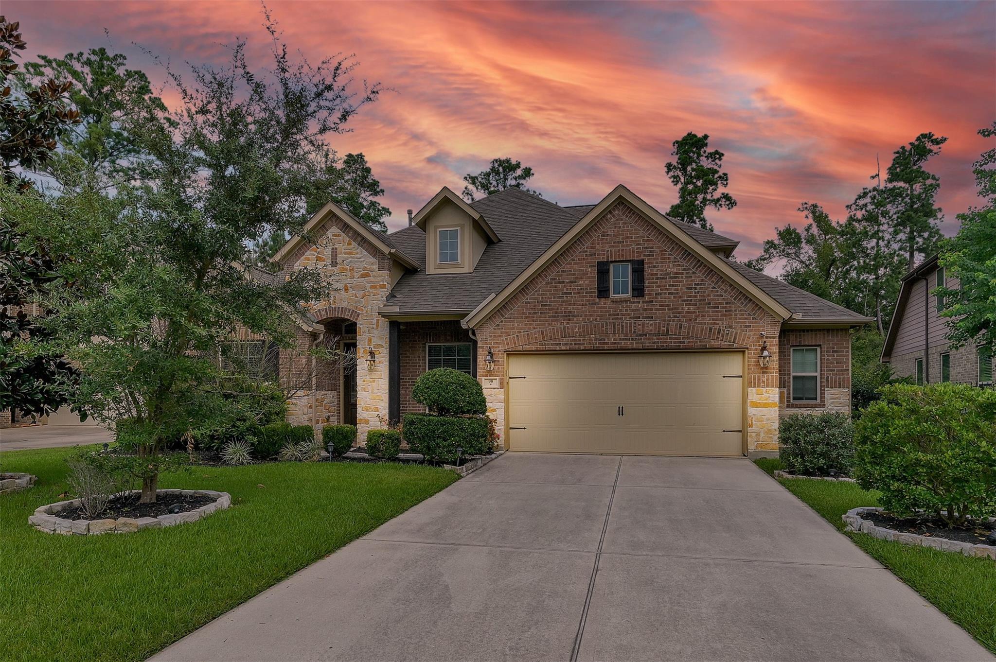 Brick-and-stone house with a two-car garage and landscaped front yard beneath a colorful dusk sky.