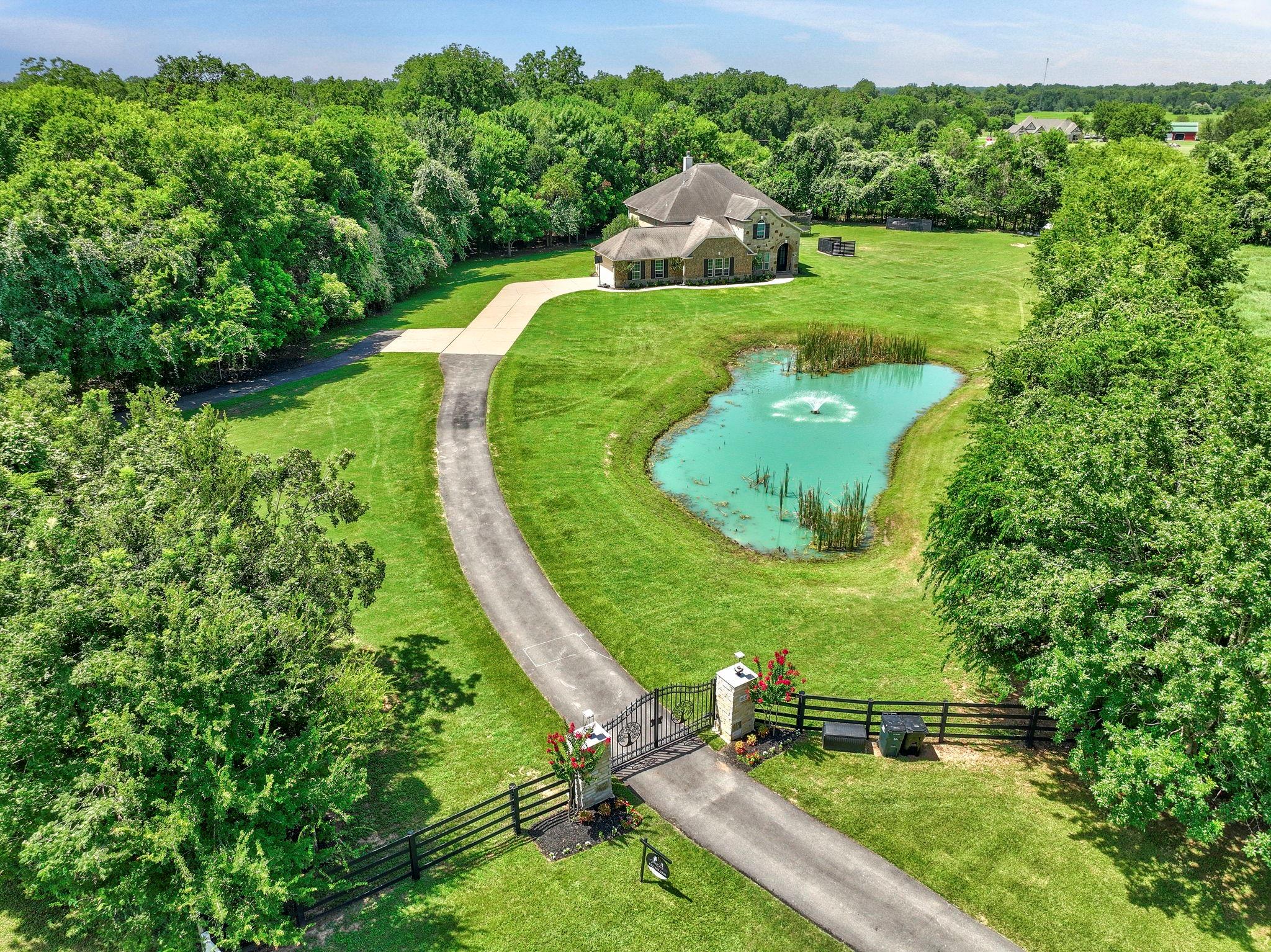 Aerial view of a brick home with a gated driveway and fountain pond on a tree-lined property.