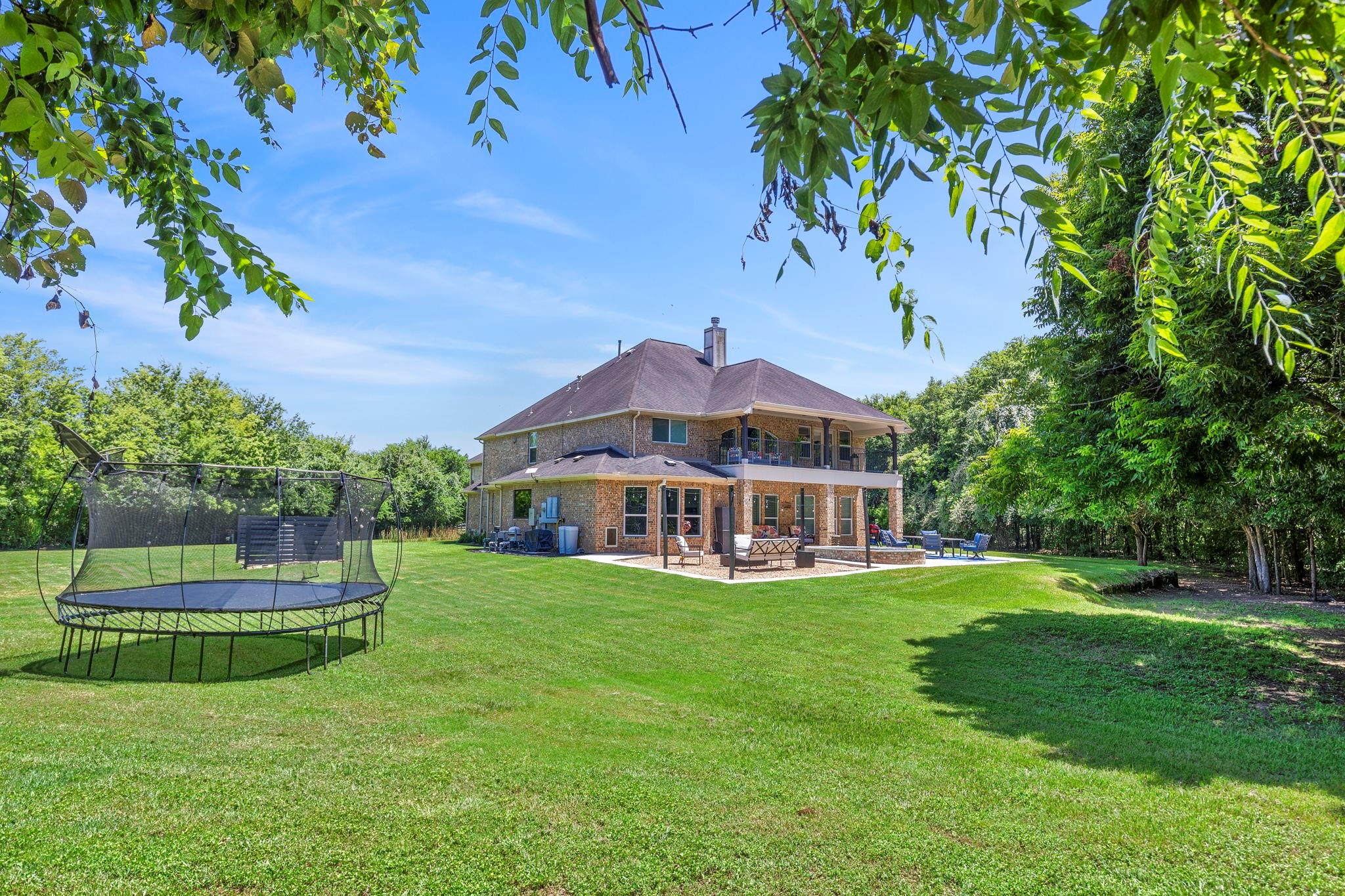 Two-story brick home with a covered patio, broad lawn, trampoline, and mature trees.