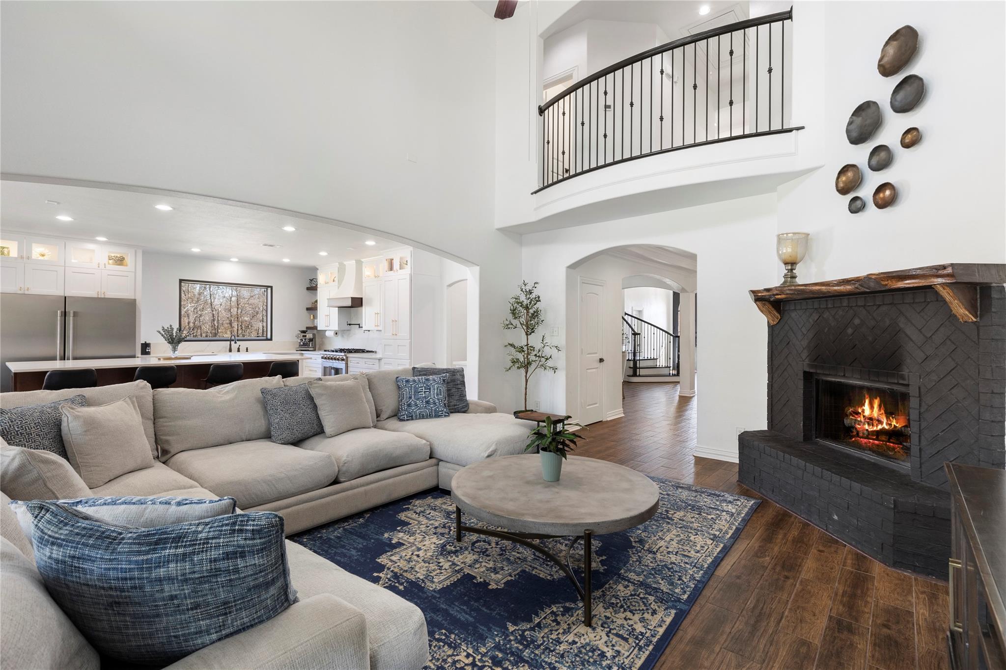 Open-plan living room with a large sectional, lit brick fireplace, and curved upstairs balcony.