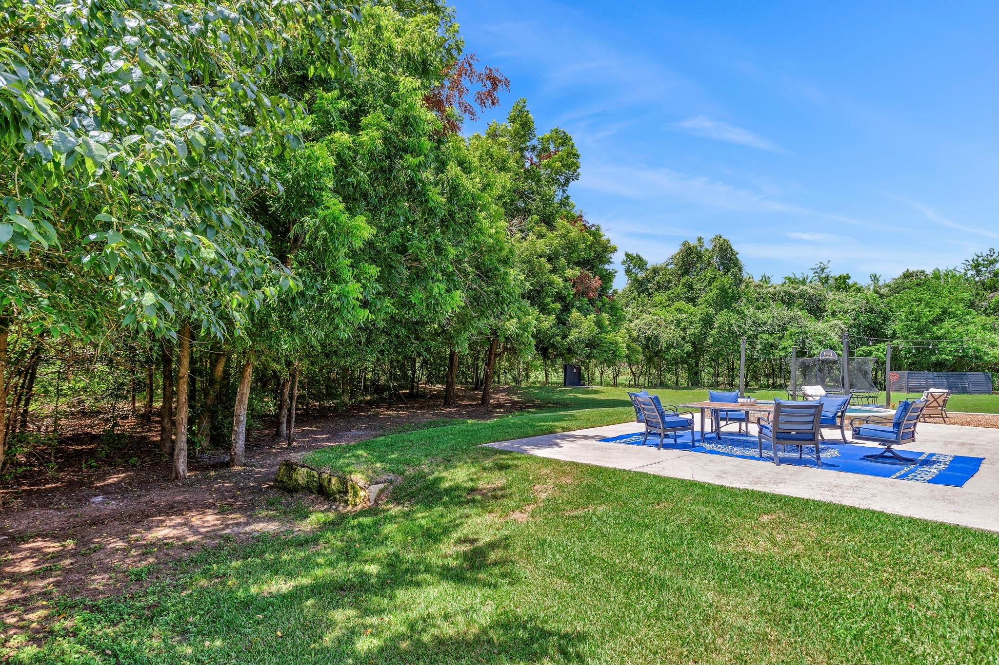 Backyard patio with blue seating, a broad lawn, mature trees, and a fenced basketball area.