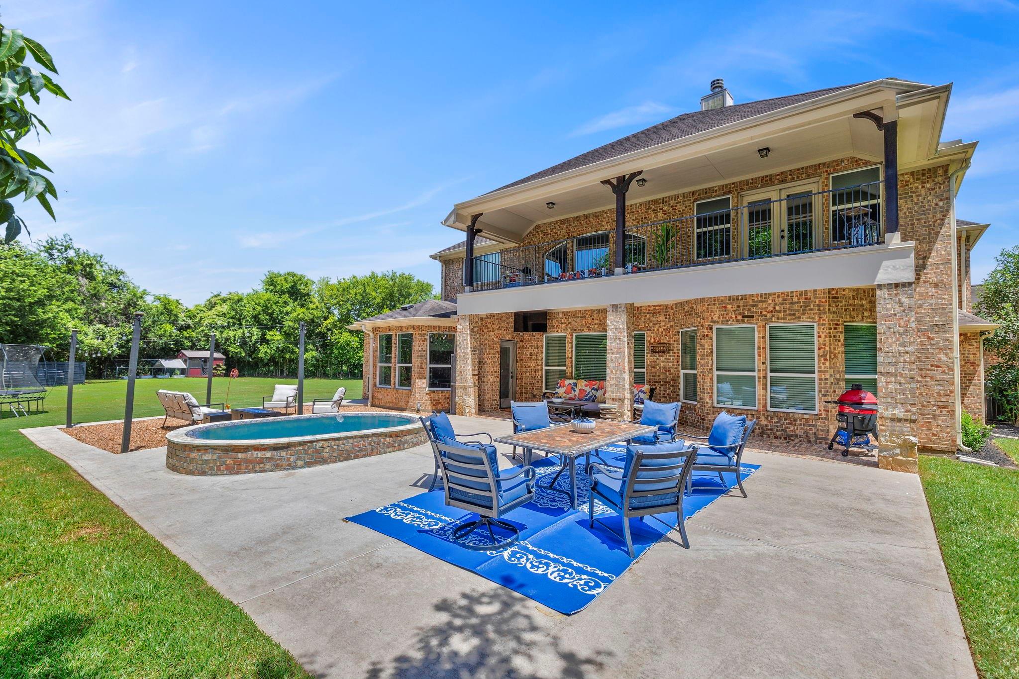 Brick home’s rear patio with a raised pool, outdoor dining set, and covered second-story balcony.