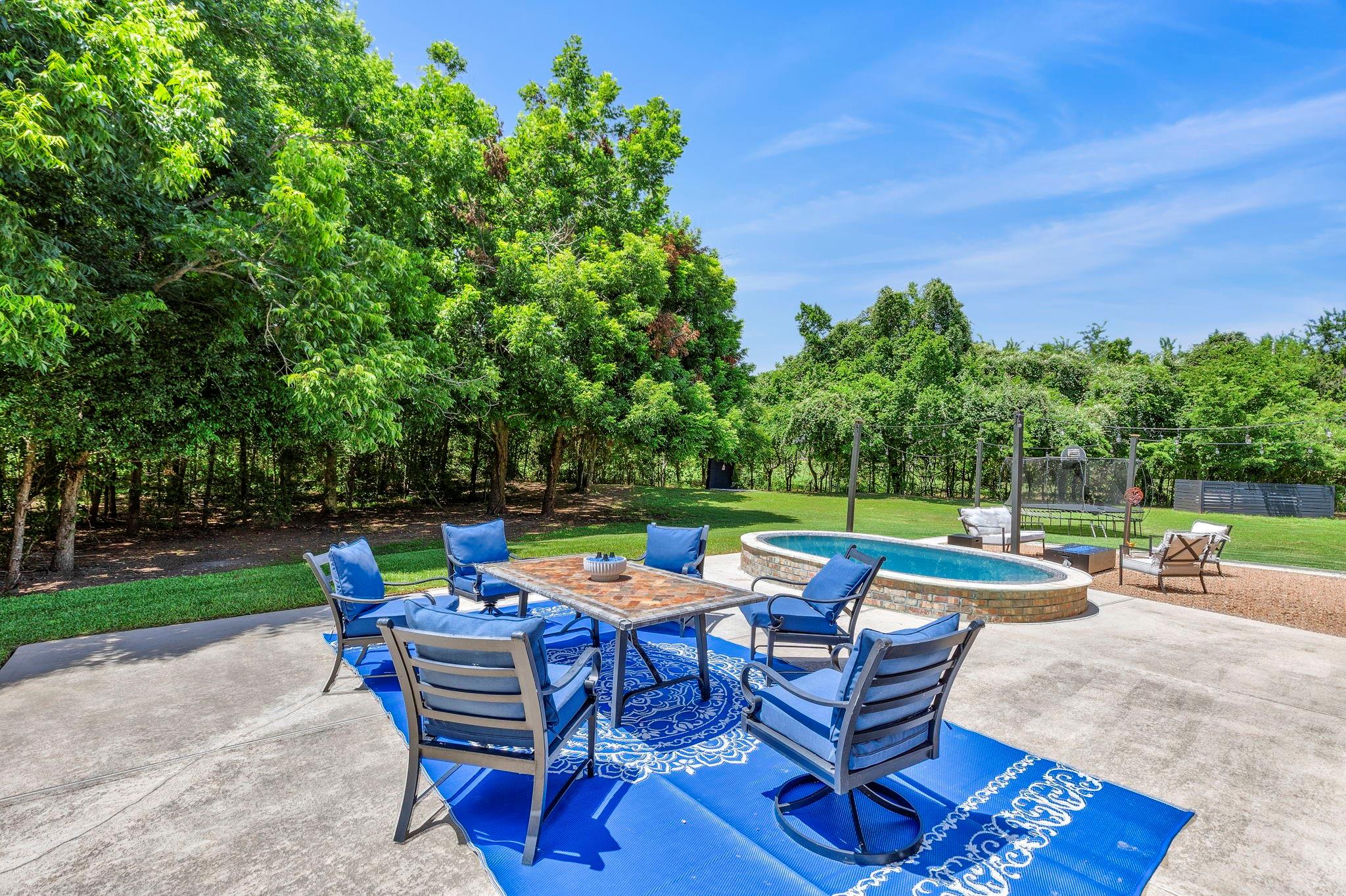 Backyard patio with a round pool and blue outdoor dining chairs beneath mature trees.