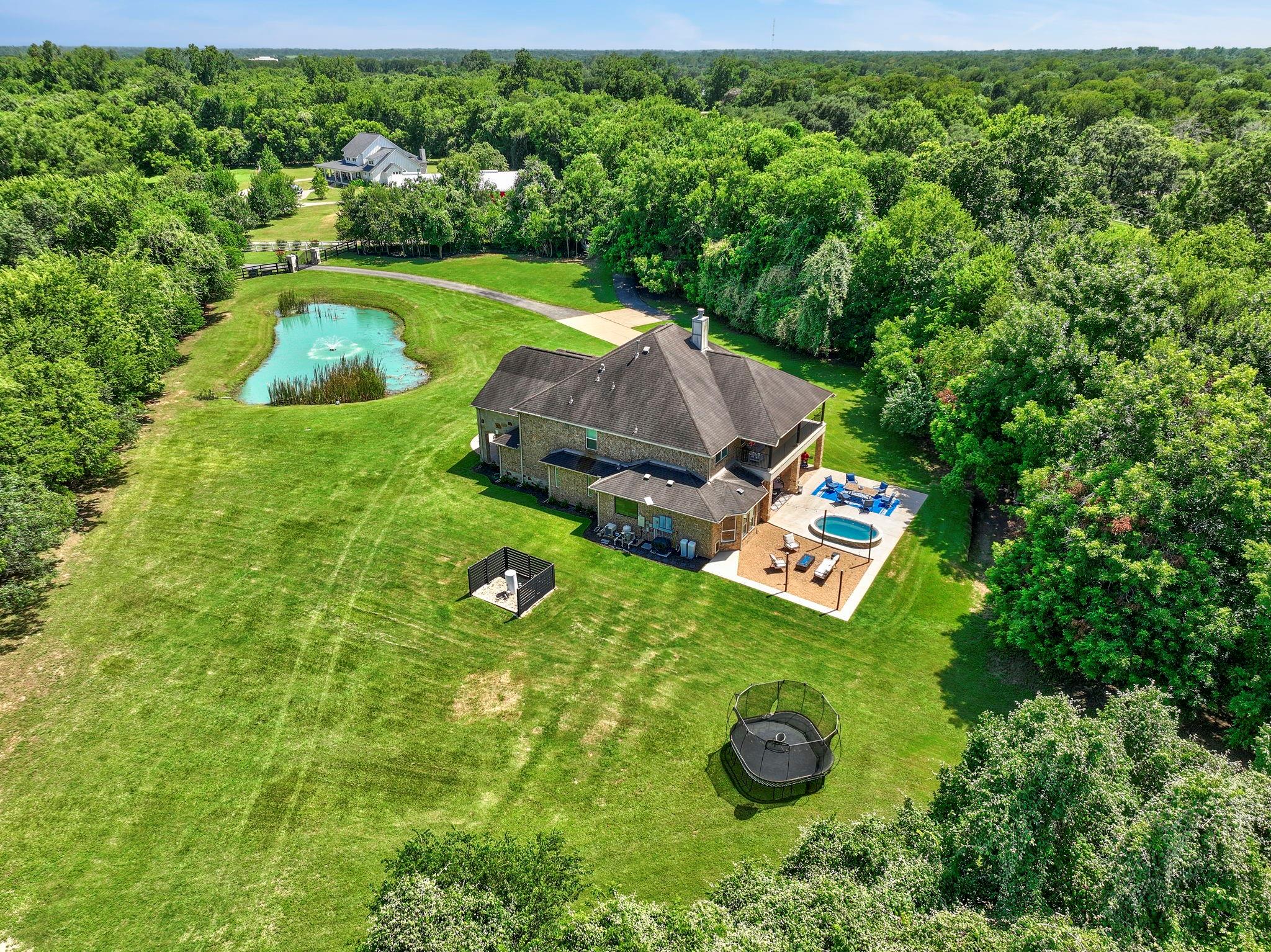 Aerial view of a home with a pool, pond, and broad tree-lined lawn.