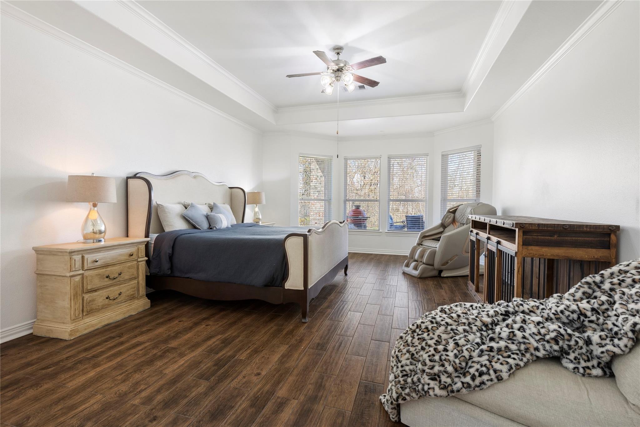 Furnished bedroom with a tray ceiling, dark wood-look flooring, massage chair, and windows with blinds.