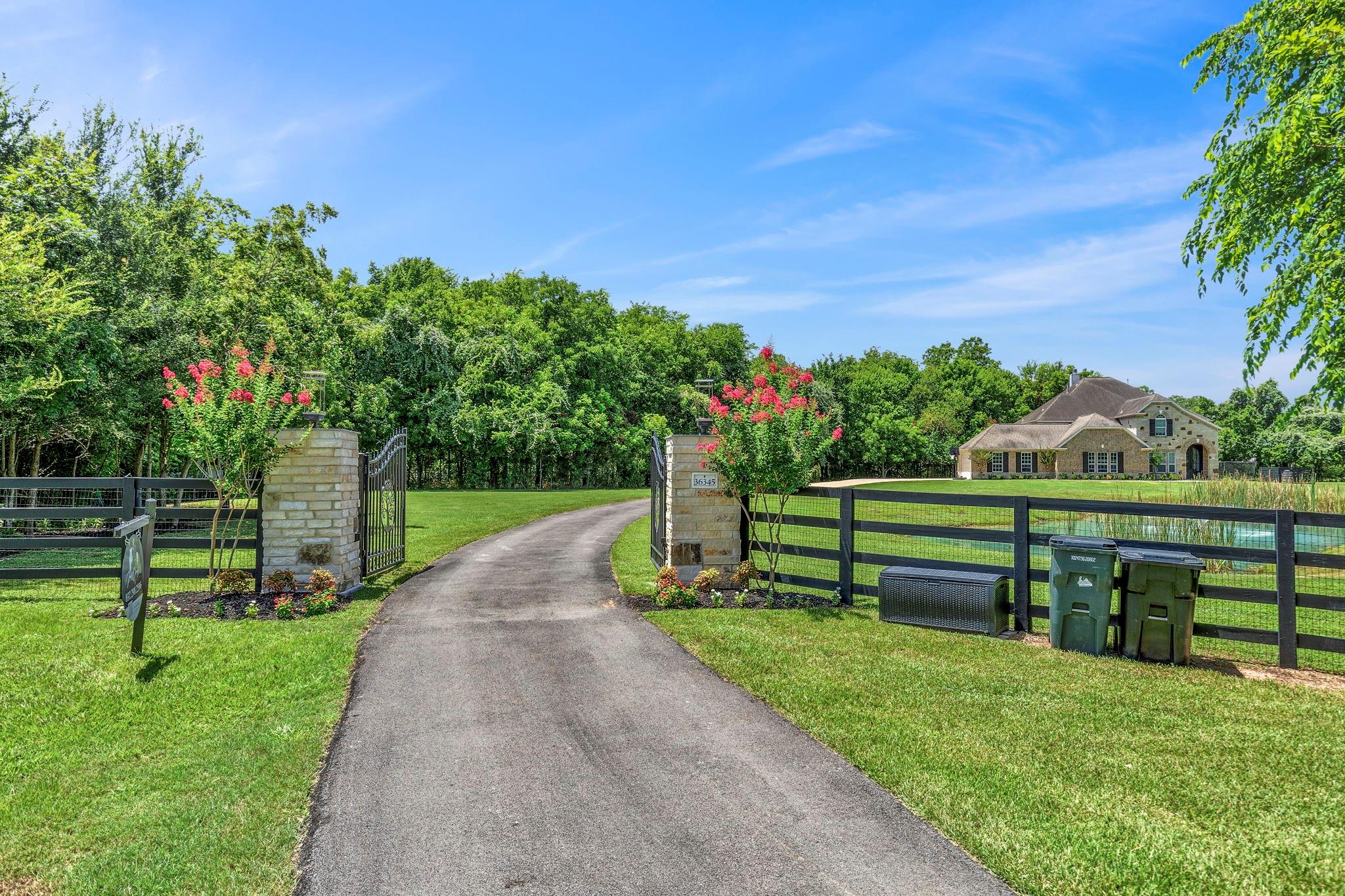 Stone house beyond a gated driveway, with black fencing, flowering trees, and a pond.