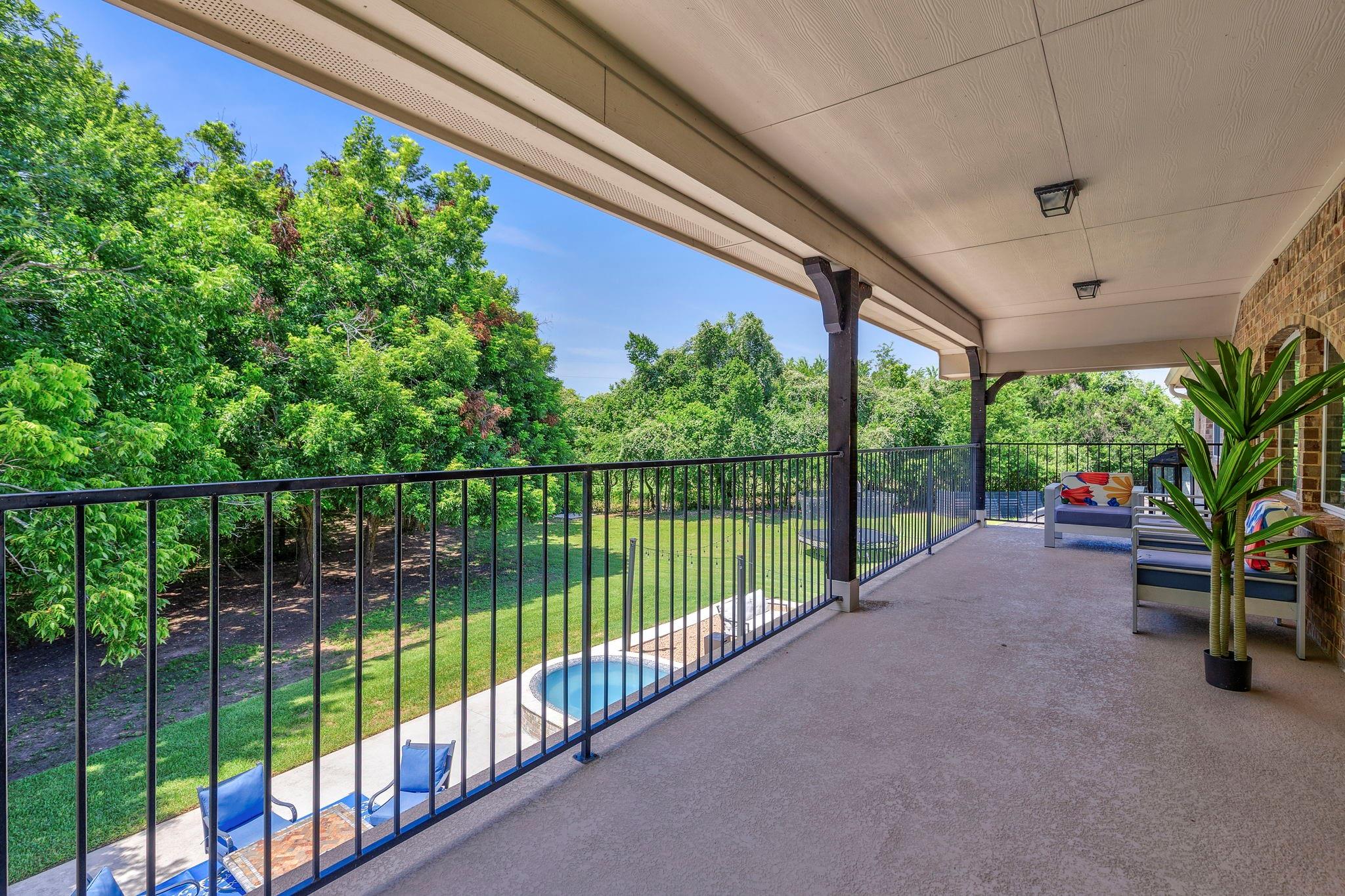 Covered patio with seating and brick arches overlooks a lawn, mature trees, and a small pool.