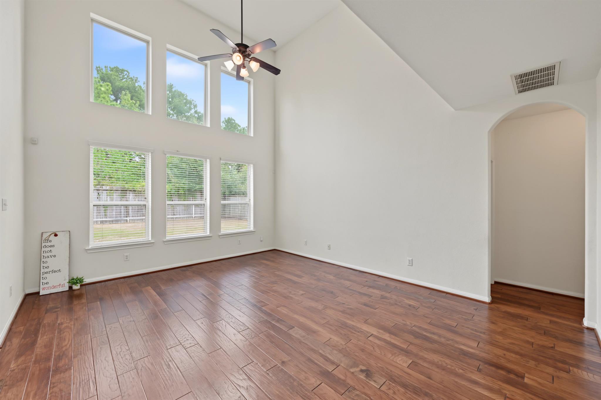 Empty living room with a vaulted ceiling, tall windows, ceiling fan, and reddish-brown wood floors.
