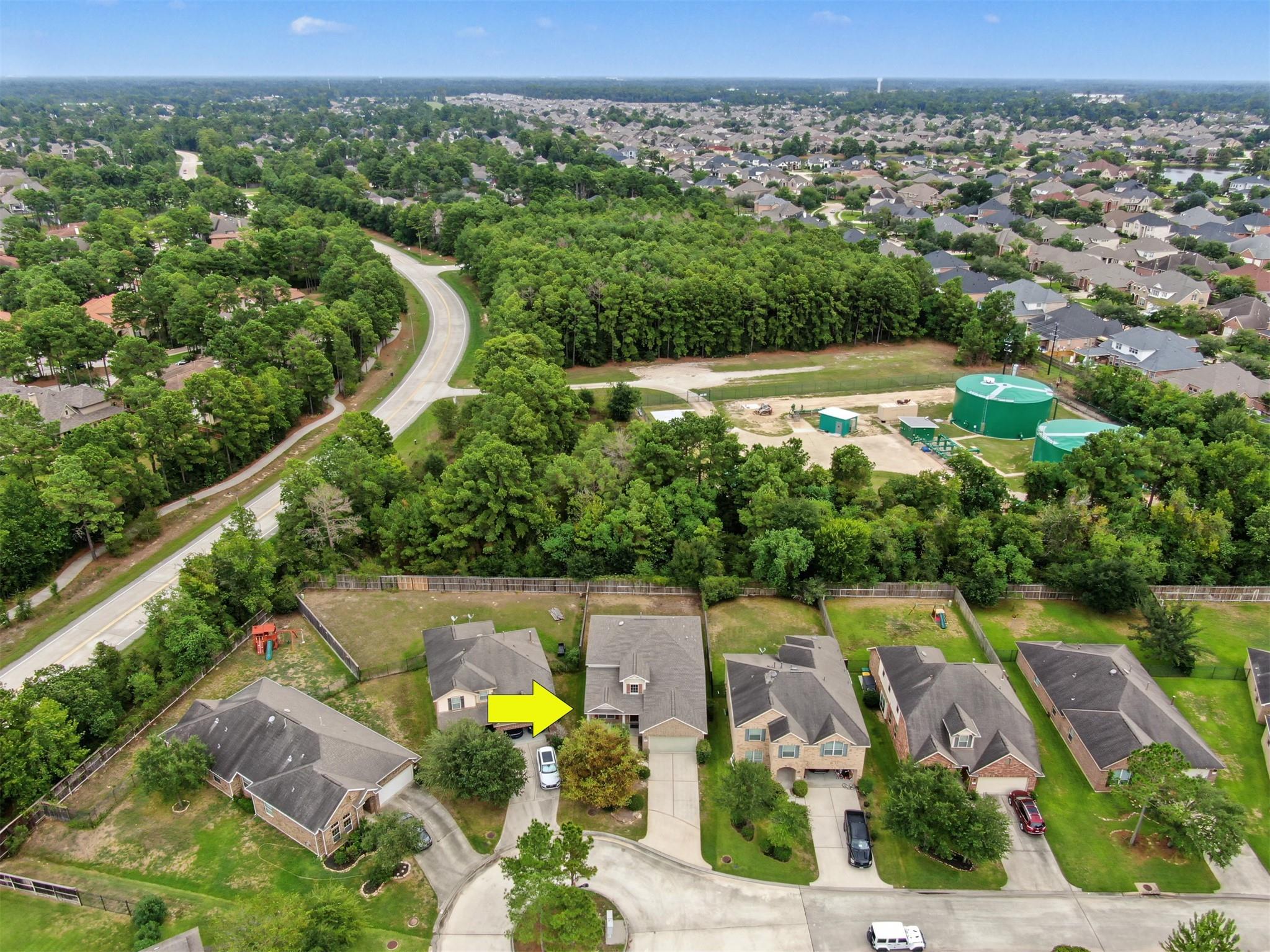 Aerial view of a two-story home marked by a yellow arrow, beside dense woods and a neighborhood road.