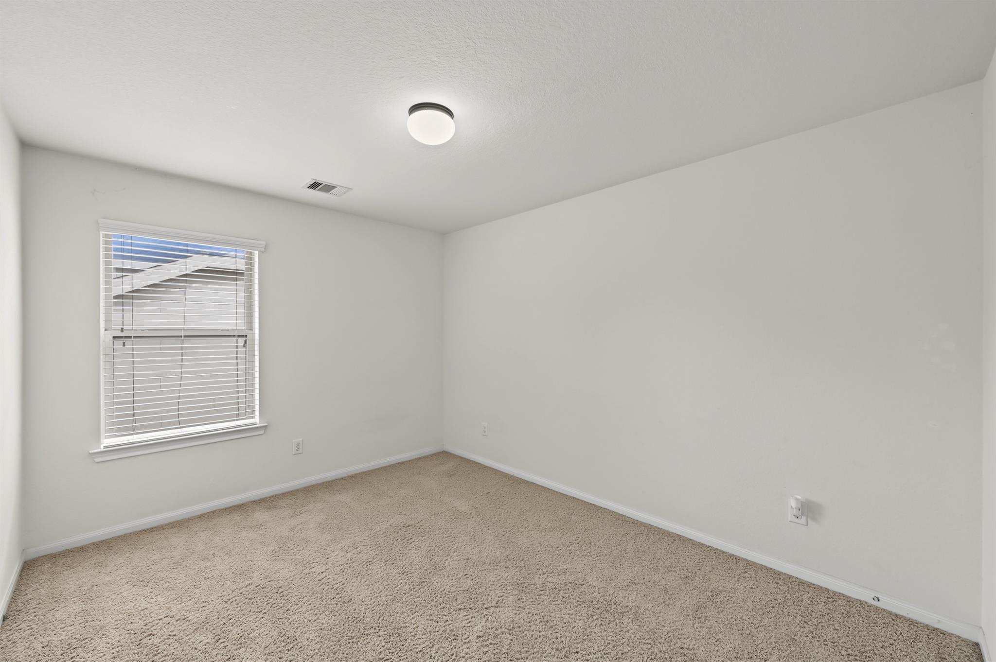 Empty carpeted room with white walls, a window with blinds, and a ceiling light.