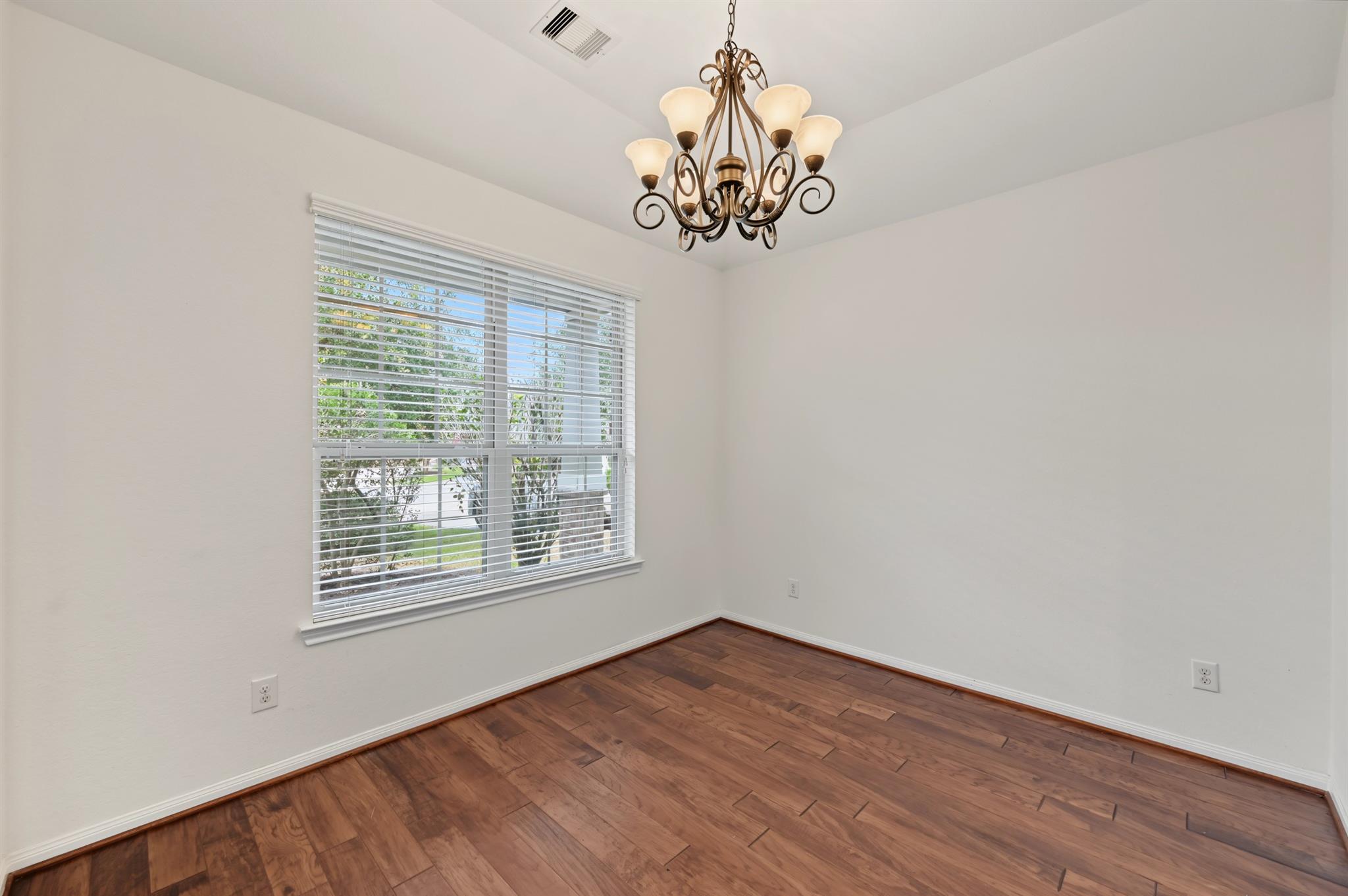 Empty room with dark hardwood floors, a wide window with blinds, and an ornate chandelier.