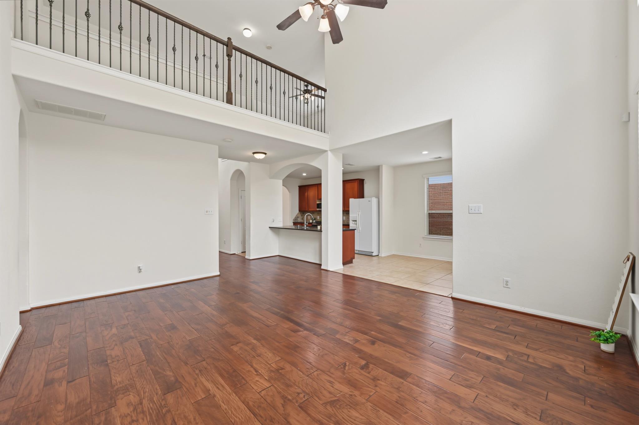 Open living room with wood floors, vaulted ceiling, upstairs railing, and kitchen beyond.