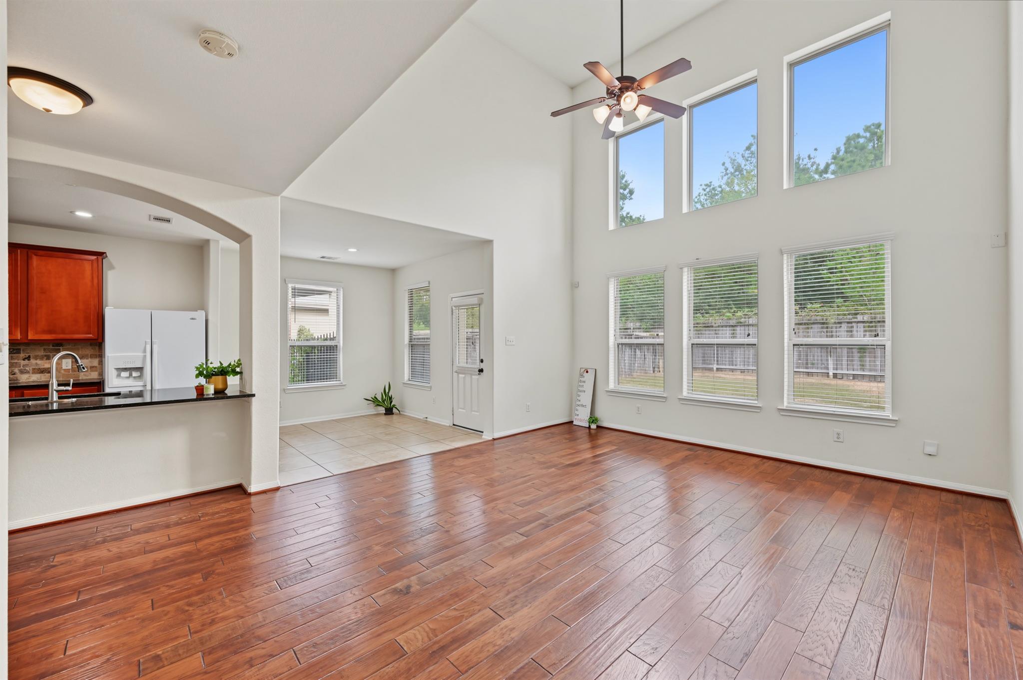 Open living area with hardwood floors, vaulted ceiling, tall windows, and an adjoining kitchen.