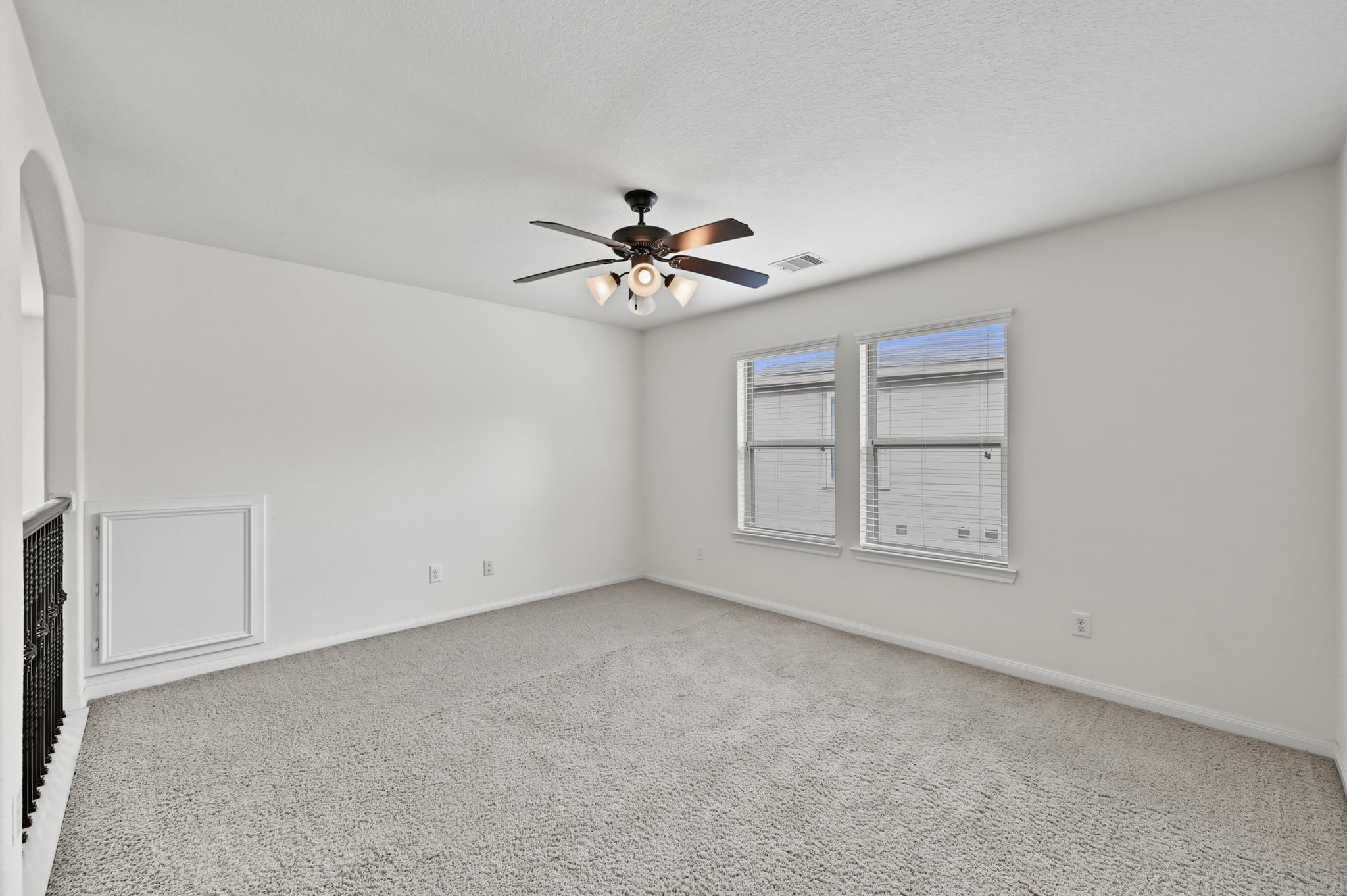 Empty carpeted room with two windows, a ceiling fan, and a stair railing.