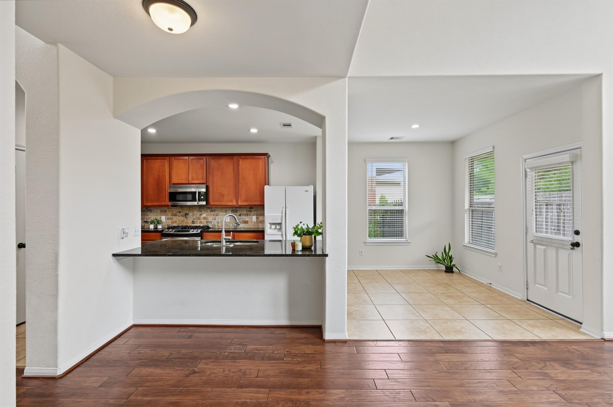 Open kitchen with wood cabinets and breakfast bar beside a tiled breakfast area with windows and a glass-paneled door.