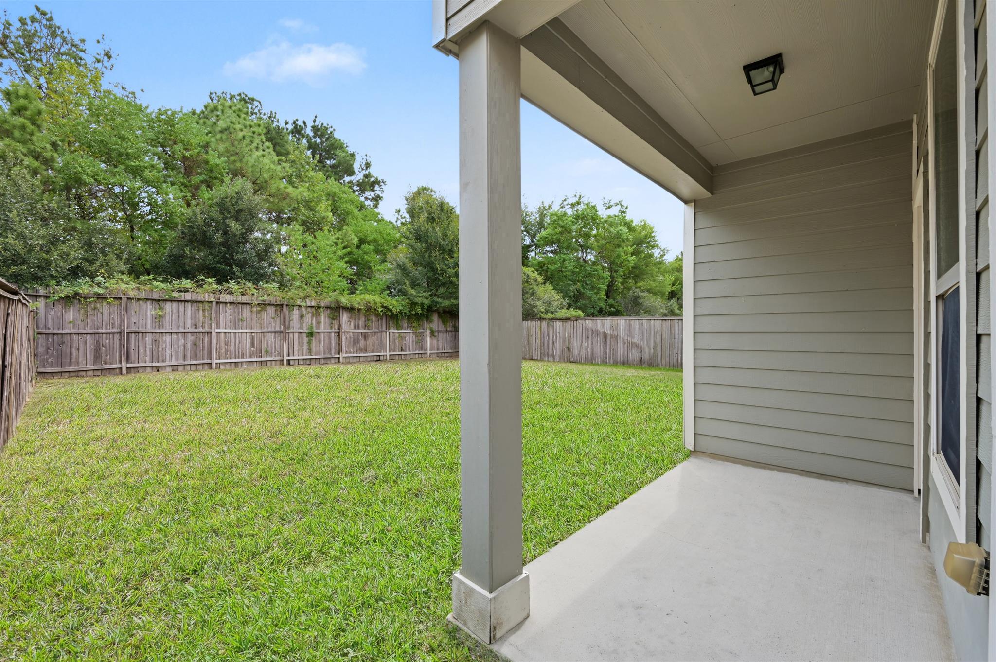 Covered concrete patio overlooking a fenced grassy backyard with mature trees.