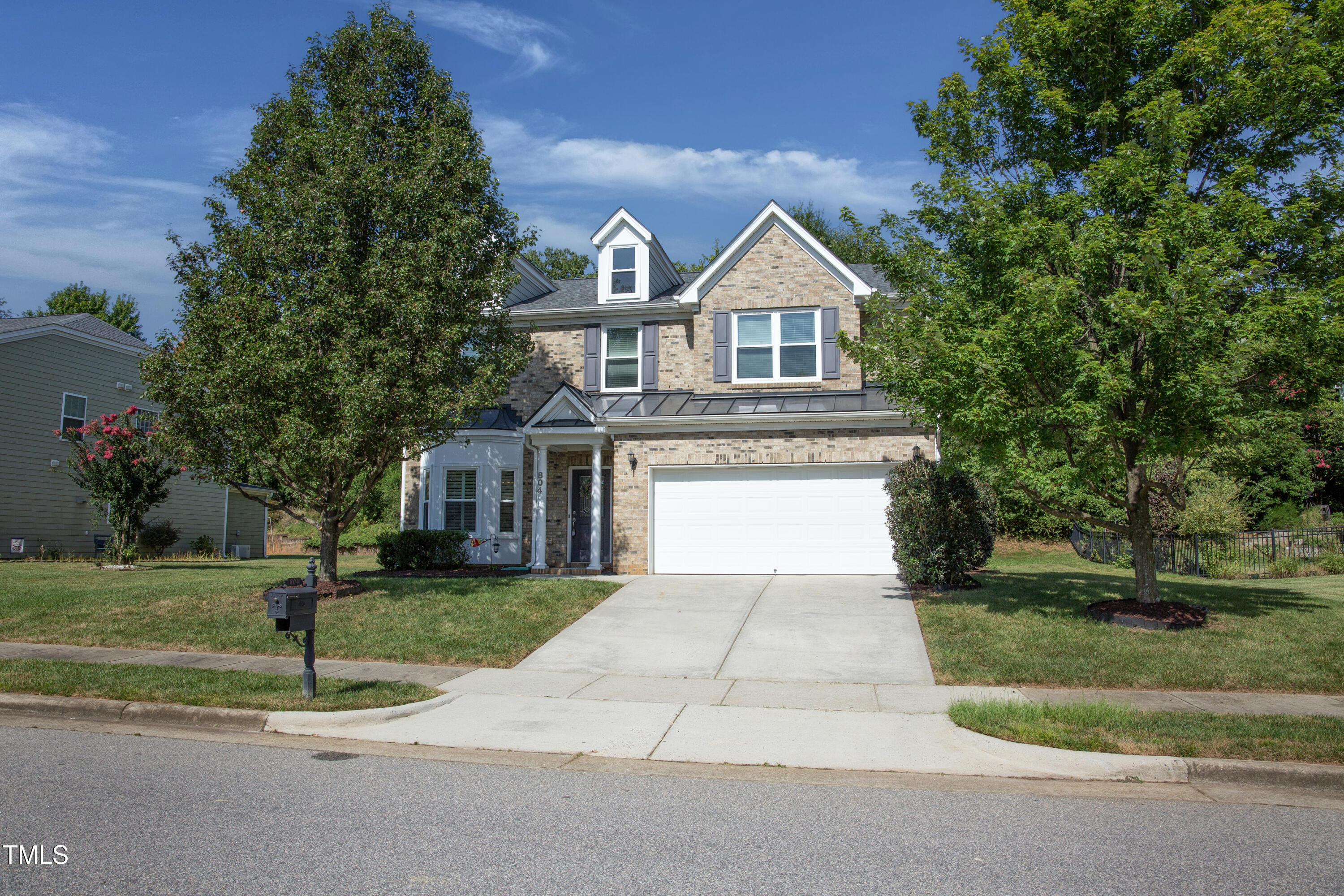 Two-story brick house with gray shutters, a white garage door, and mature trees framing the lawn.